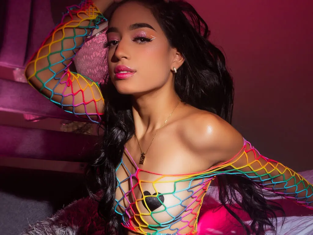 AbbyNears poses in a colorful rainbow fishnet top that shows off her chest and cleavage against a dark background.