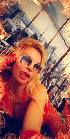 Blonde woman in a red outfit and sunglasses posing against a city skyline with a fiery overlay effect.
