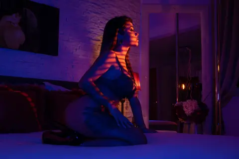 Sensual woman in black lingerie posing on a bed in a moody, purple-lit bedroom setting.