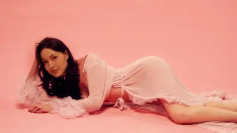 Young woman in a white flowing lace dress lying on a pink background in a dreamy, romantic mood.