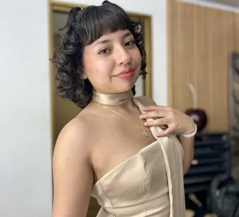 Smiling young woman in a beige halter neck dress posing in a bright, minimalist indoor setting.