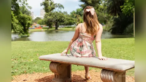 Young woman in a floral summer dress sitting on a stone bench by a peaceful lake in a sunny park.