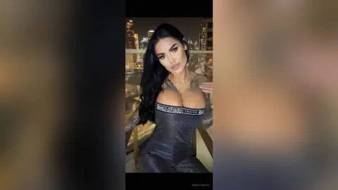 Glamorous brunette woman in a tight grey bodysuit posing on a city balcony at night.