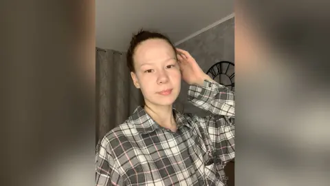 Young woman in a grey plaid flannel shirt posing in a cozy indoor setting with a natural, relaxed mood.