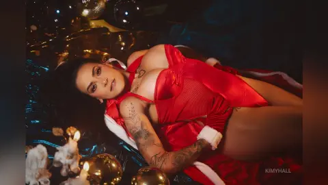 Tattooed woman in a red and white festive bodysuit lying on shimmering blue fabric for a holiday mood.