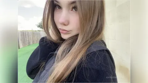 Young woman with long brown hair wearing a black hoodie in a bright outdoor setting.