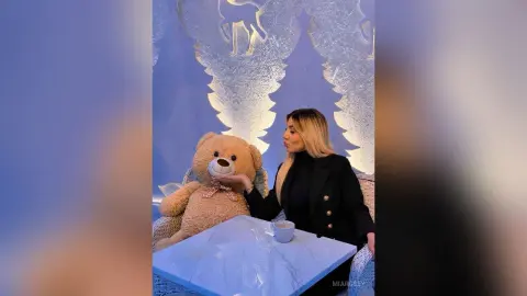 Blonde woman in a black blazer sitting at a marble table with a large teddy bear in a dreamy blue room.