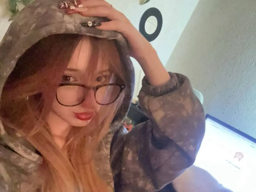 Young woman with long blonde hair and glasses wearing a cozy grey camouflage hoodie in a dimly lit room.