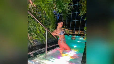 NicoleBella poses in a black and white patterned bikini while standing in a lit swimming pool surrounded by tropical palm leaves.