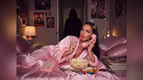 Woman in pink silk pajamas relaxing on a bed with popcorn and a vintage pink telephone in a cozy bedroom.