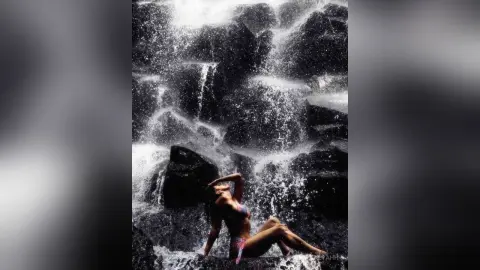 Aalyahh poses naked under a splashing waterfall, sitting on dark rocks with her arms raised above her head.
