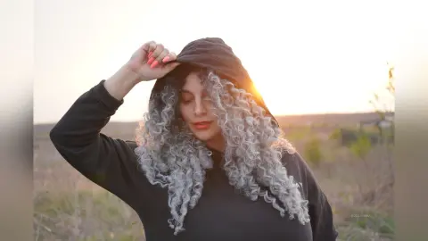 Woman with long curly silver hair wearing a black hoodie in a sunlit field at sunset.