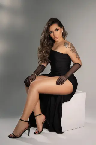 EvaSalvatore poses in a black strapless dress and sheer gloves, showing off her long legs and shoulder tattoo while sitting on a white block.