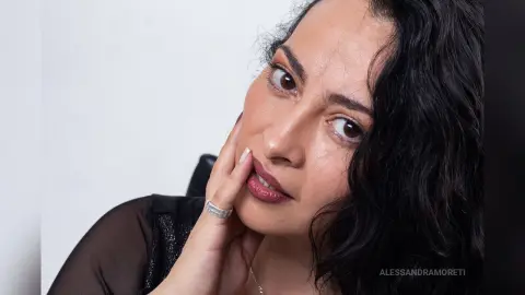 Close-up portrait of a woman with wavy dark hair and dark lipstick posing in a black sheer outfit.