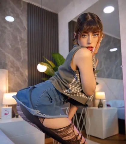 Young woman in a grey knit tank top and denim shorts posing in a modern, brightly lit bedroom.