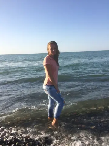 Young woman in a pink polo shirt and blue jeans standing on a rocky beach by the ocean under a clear sky.