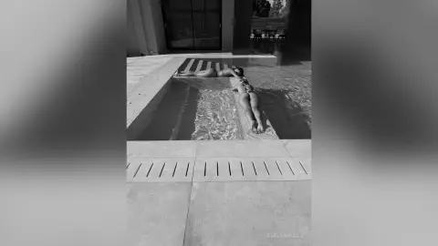 EvelynMills lies naked on the edge of a swimming pool, showing her bare back and buttocks while partially submerged in the water.