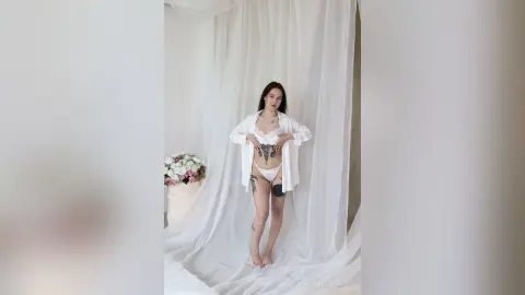 Tattooed woman in white lingerie and open shirt posing in a bright, ethereal studio with white drapery.