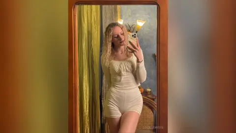 Blonde woman in a white long-sleeve bodysuit taking a mirror selfie in a cozy, sunlit room.