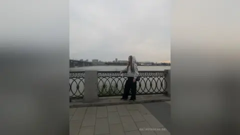 Young woman in a white blouse and black trousers leaning on a bridge railing against a cloudy city skyline.
