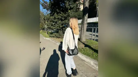 Blonde woman in a white blouse and light trousers walking on a sunny park path.