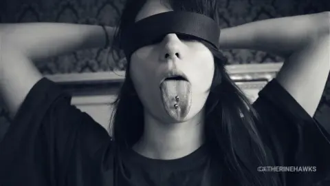 Blindfolded woman in a black t-shirt sticking out her tongue in a moody, black and white close-up portrait.