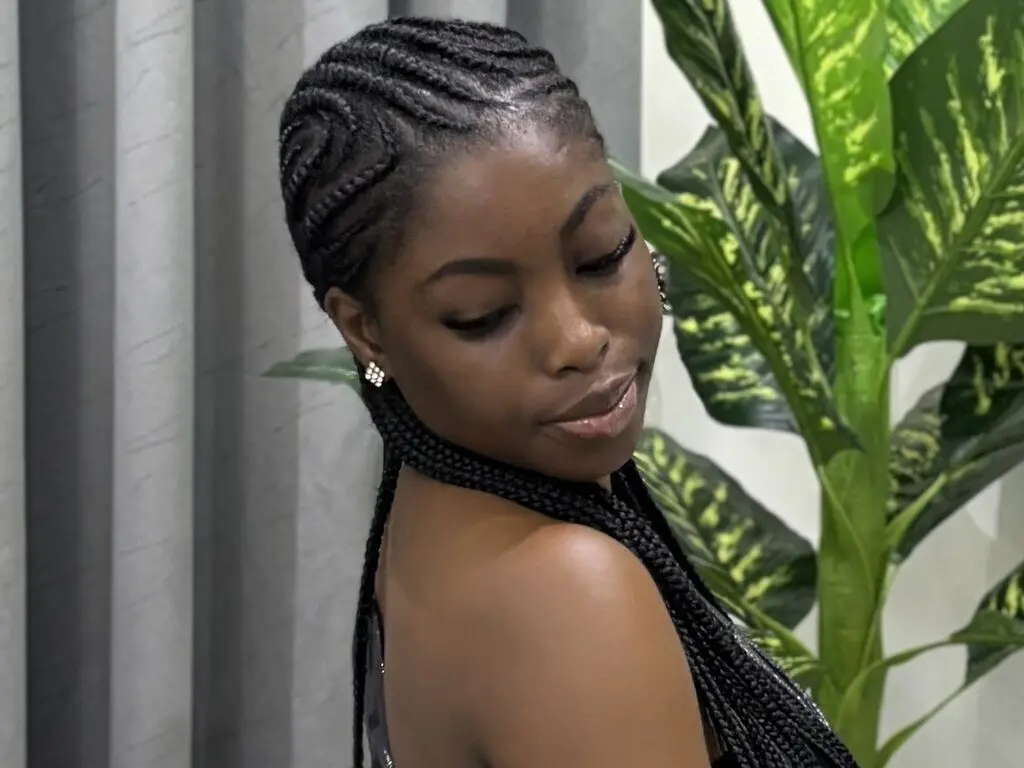 Young woman with elegant black cornrow braids posing serenely near lush green indoor plants.