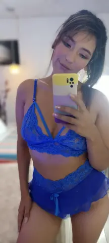 Young woman in blue lace lingerie set taking a mirror selfie in a bright bedroom.