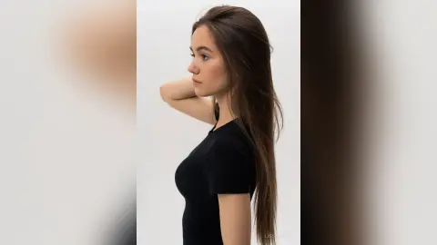 LissaDesso poses in profile wearing a tight black t-shirt with her long brown hair flowing down her back.