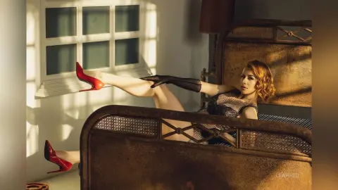 Elegant woman in a black mesh dress and red high heels lounging on a vintage bed in warm sunlight.