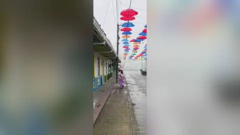 Woman in a pink dress walking under colorful umbrellas on a wet city street during a rainy day.