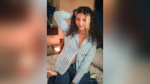 Young woman with curly hair in a blue striped shirt relaxing on a bed in a cozy, warm-lit bedroom.