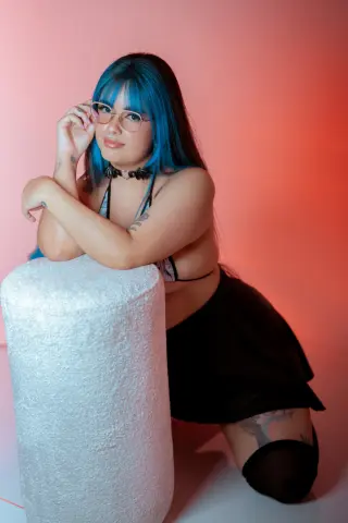 Woman with blue hair and glasses wearing a black outfit posing against a pink studio background.