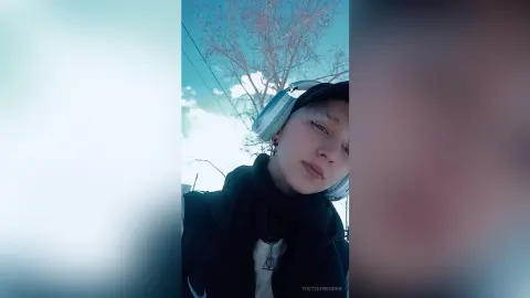Young woman with light blue headphones and black winter jacket relaxing outdoors under a bright sky.
