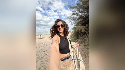 SophieeSunsett — SophieeSunsett smiles in sunglasses and a black tank top while standing on a sandy beach near the ocean.