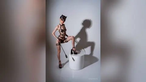 Fashion model in black strappy bodysuit and mask posing on a white pedestal against a minimalist gray background.