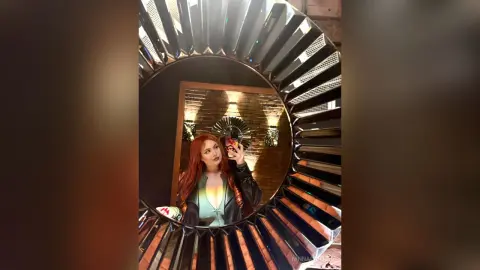 Redheaded woman in a black leather jacket taking a selfie in a decorative sunburst mirror with warm lighting.