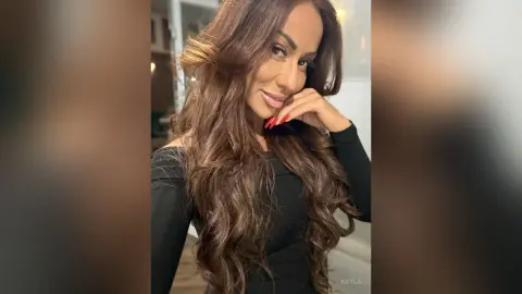 Beautiful woman with long wavy brown hair wearing a black long-sleeve top in a soft indoor setting.