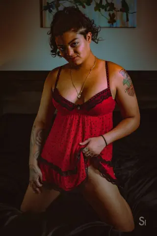 Young woman in a red polka dot lace chemise sitting on a dark bed in a moody, intimate bedroom setting.