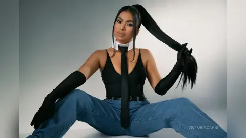 Stylish woman in a black bodysuit, necktie, and long gloves sitting in a modern studio setting.