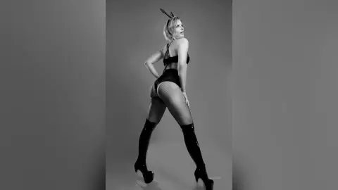 Gia poses in black lingerie, fishnet stockings, and thigh-high boots against a plain background.