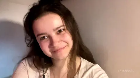 Smiling young woman with long brown hair wearing a casual cream ribbed top in a soft indoor setting.