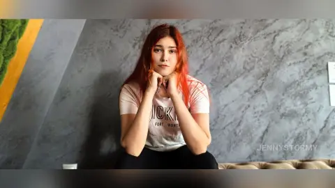 Red-haired woman in a white graphic t-shirt sitting thoughtfully against a grey marble wall background.