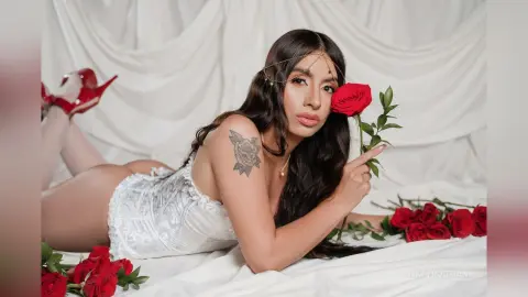 Glamorous woman in white lace lingerie lying on white sheets holding a red rose in a romantic setting.