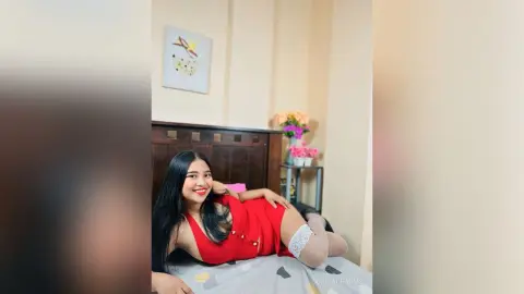 Smiling young woman in a red dress and white stockings relaxing on a bed in a bright bedroom.