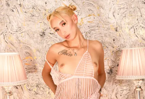 Blonde woman in pink sheer lingerie posing against a marble background with elegant lamps.