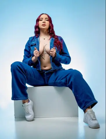 Red-haired woman in denim jacket and jeans sitting on a white box against a blue studio background.