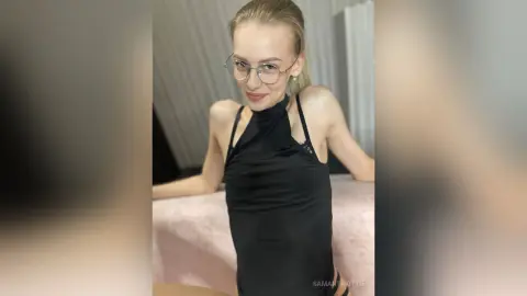 Smiling blonde woman in a chic black halter neck top posing in a bright, minimalist indoor setting.