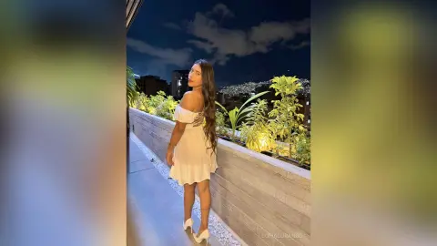 Woman in a white off-the-shoulder mini dress posing on a balcony overlooking a city skyline at night.