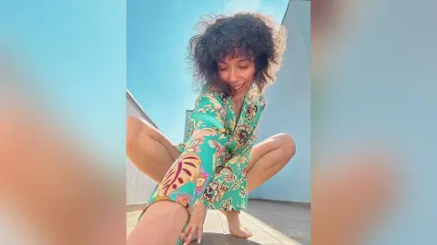 Smiling woman in a colorful floral print robe sitting outdoors under a bright blue sky.